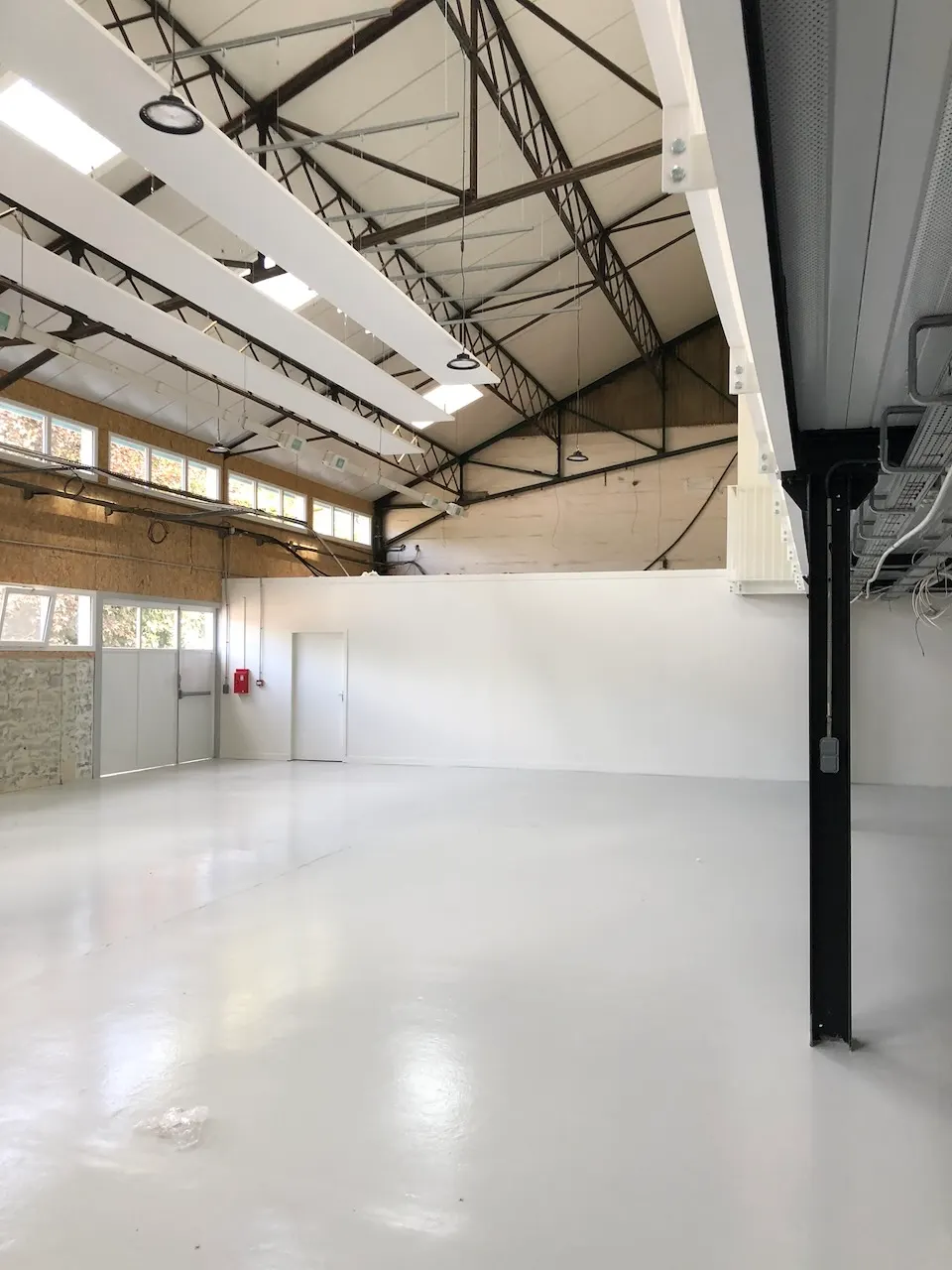 Grand volume clair sous charpente métallique, sol blanc lisse et poteau métallique noir au premier plan.