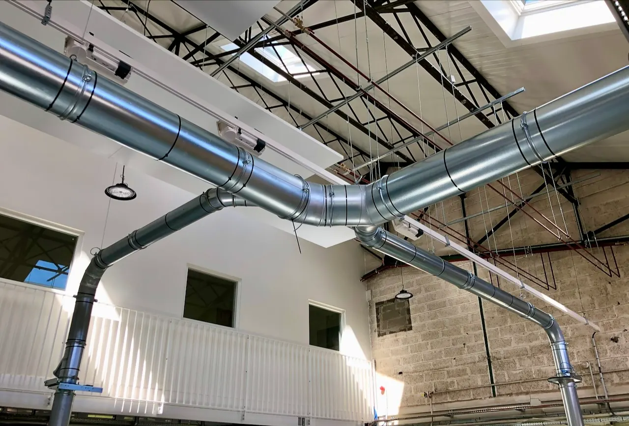 Conduits en acier galvanisé suspendus sous une charpente métallique, au-dessus de cloisons blanches percées de fenêtres.