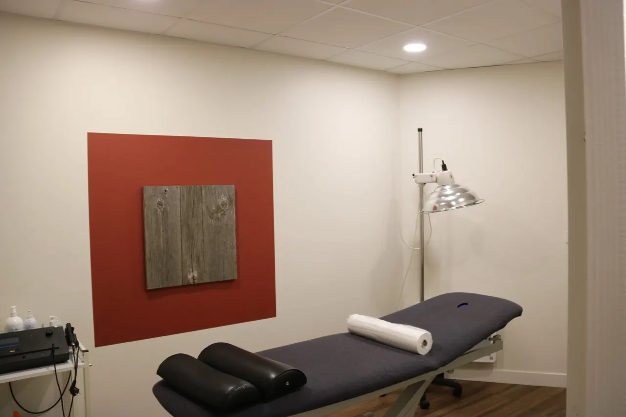 Salle de soins aux murs clairs, avec une table de soins, une lampe sur pied et un aplat rouge portant un panneau de bois.
