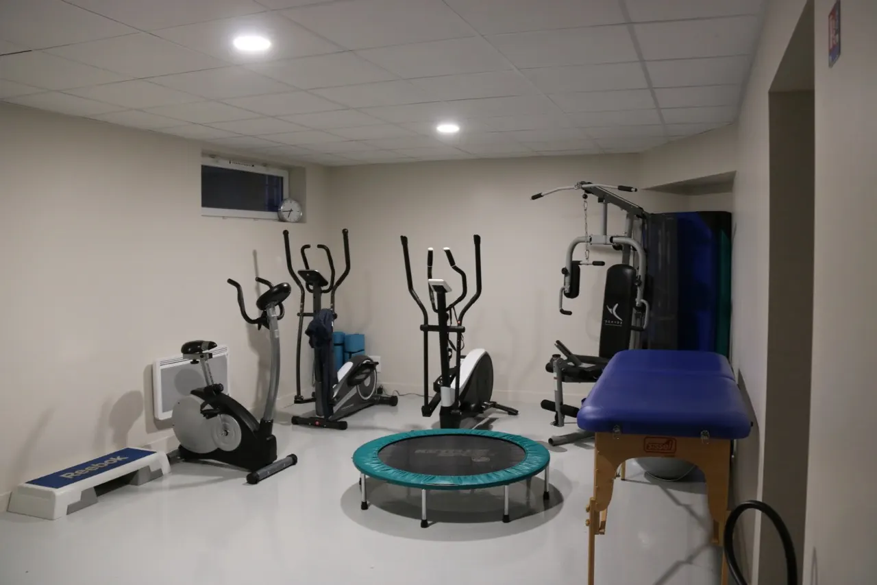 Salle claire équipée d'appareils d'exercice, d'un trampoline et d'une table de soins bleue.