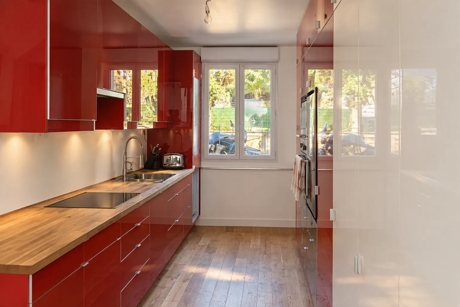 Cuisine en longueur aux façades laquées rouges et plan de travail en bois, éclairée par deux fenêtres, sur un parquet.