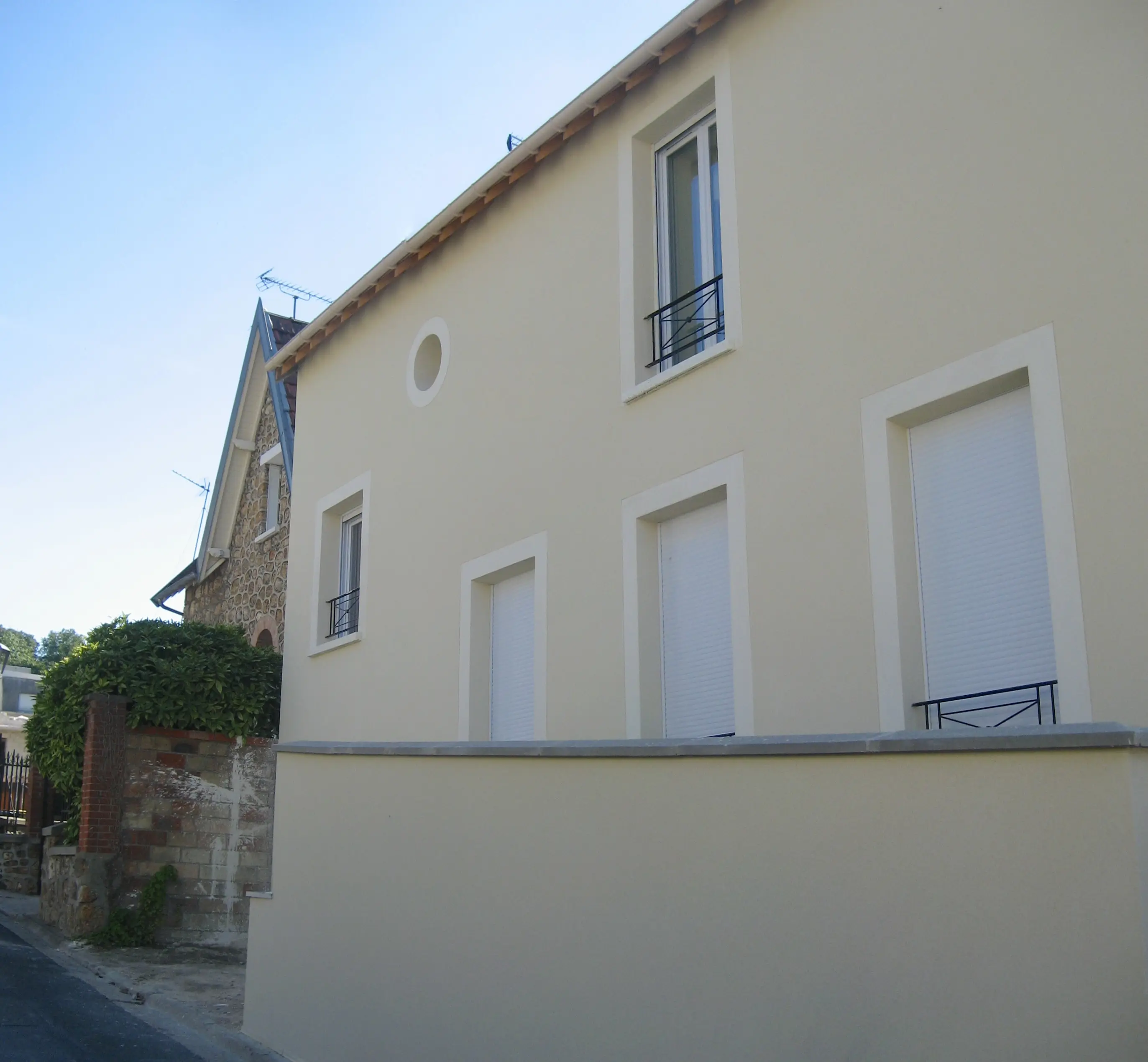 Façade enduite beige percée de fenêtres à encadrement clair et d'un oculus, au-dessus d'un mur de soubassement, à côté d'une maison voisine en meulière.