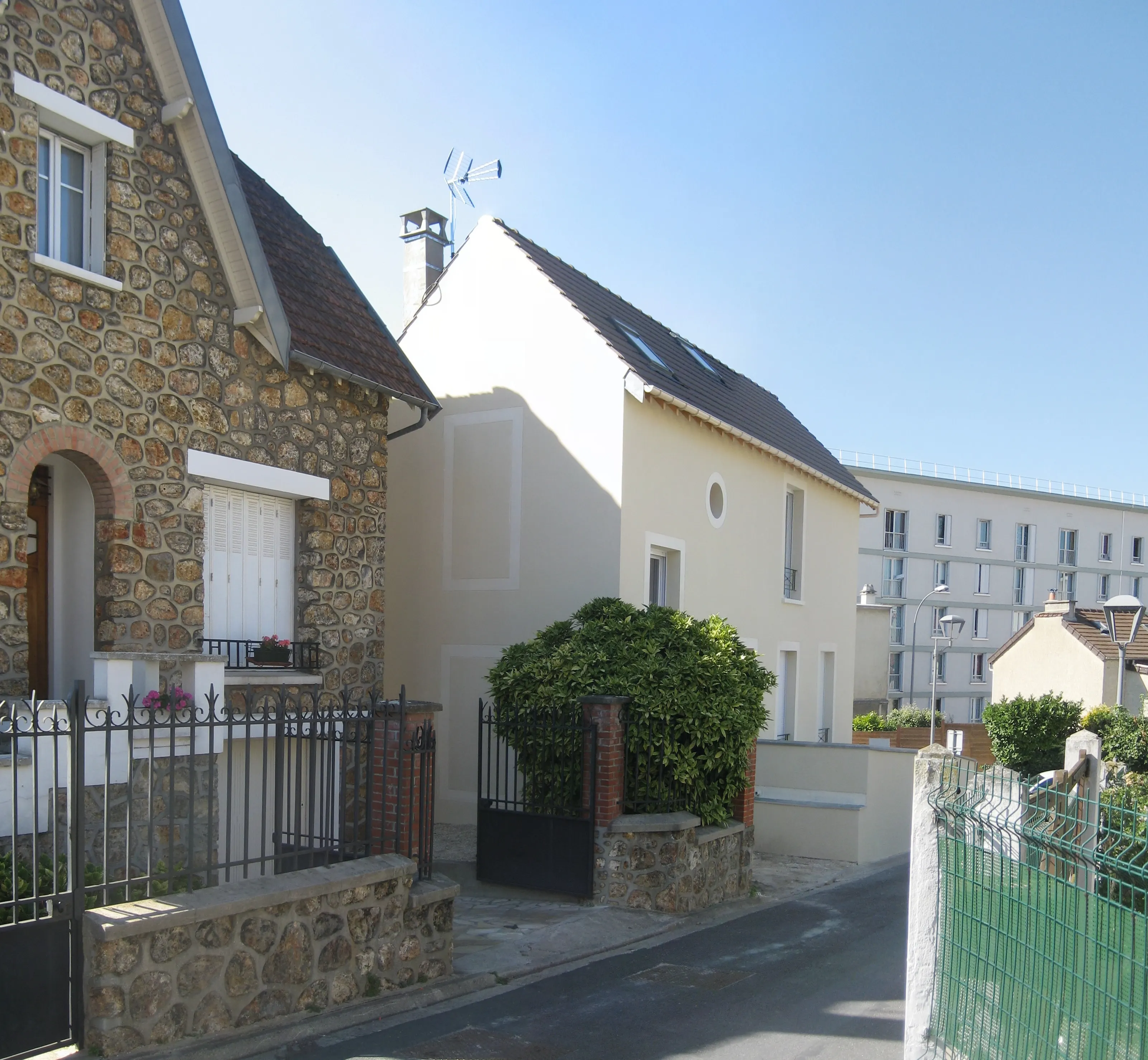 Vue de rue : à gauche une maison en meulière, au centre une maison enduite claire à toit de tuiles sombres percé de fenêtres de toit, à droite un immeuble blanc.