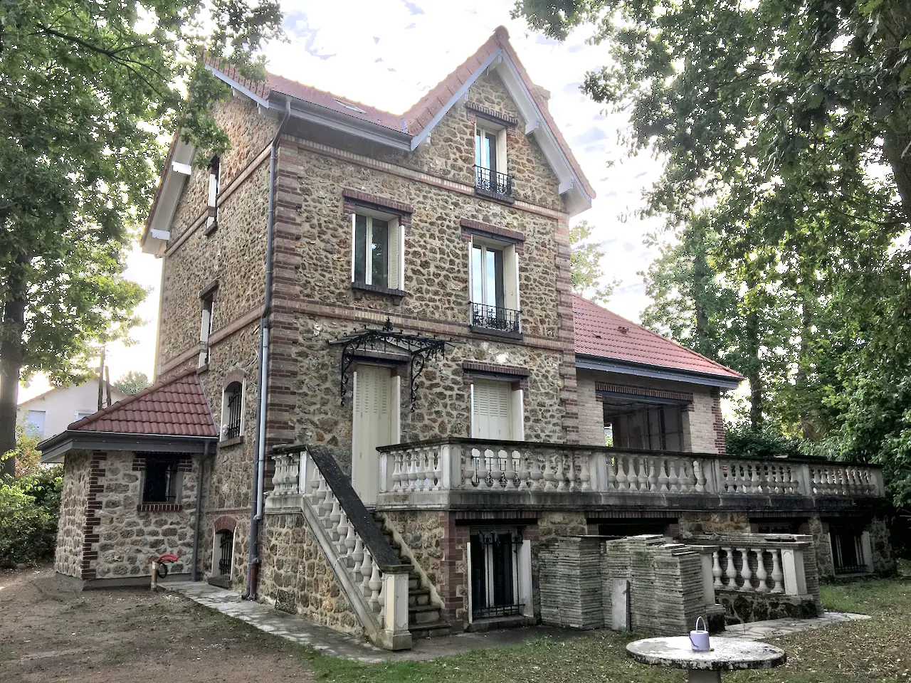 Grande maison en meulière à deux étages et toit de tuiles, avec terrasse à balustres et escalier extérieur, vue depuis un jardin arboré.