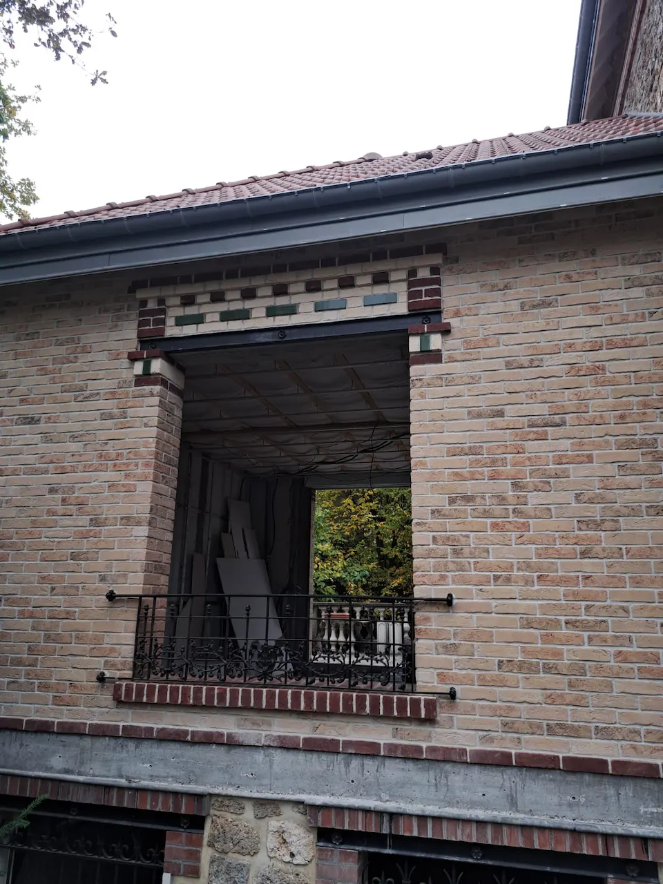 Façade en brique avec une ouverture à garde-corps en fer forgé, surmontée d'une frise décorative et d'un débord de toit.