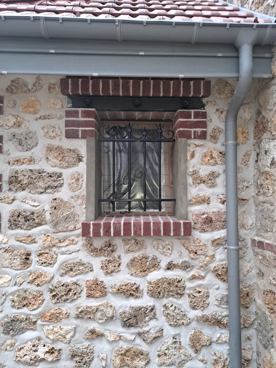 Mur en pierre meulière aux joints clairs, percé d'une petite fenêtre à grille encadrée de briques rouges, avec une descente d'eau pluviale.