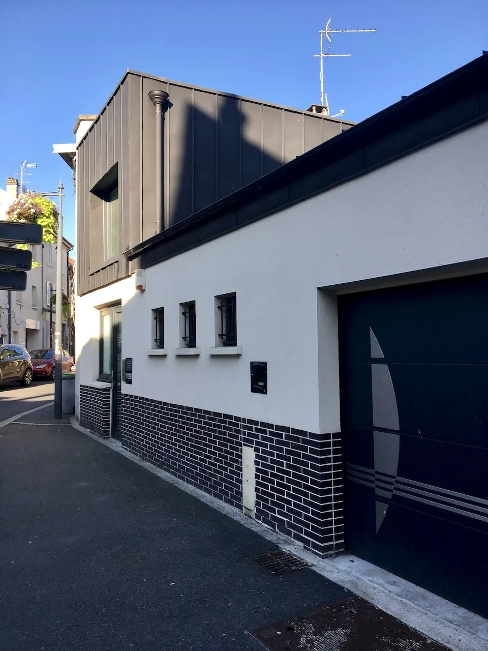 Façade latérale étroite sur un trottoir, avec un soubassement de carreaux sombres, de petites fenêtres alignées et une porte de garage foncée.