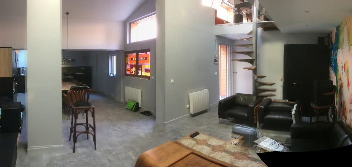 Vue panoramique d'un séjour clair sous rampant, avec poteau central, canapés sombres et escalier en colimaçon au fond.