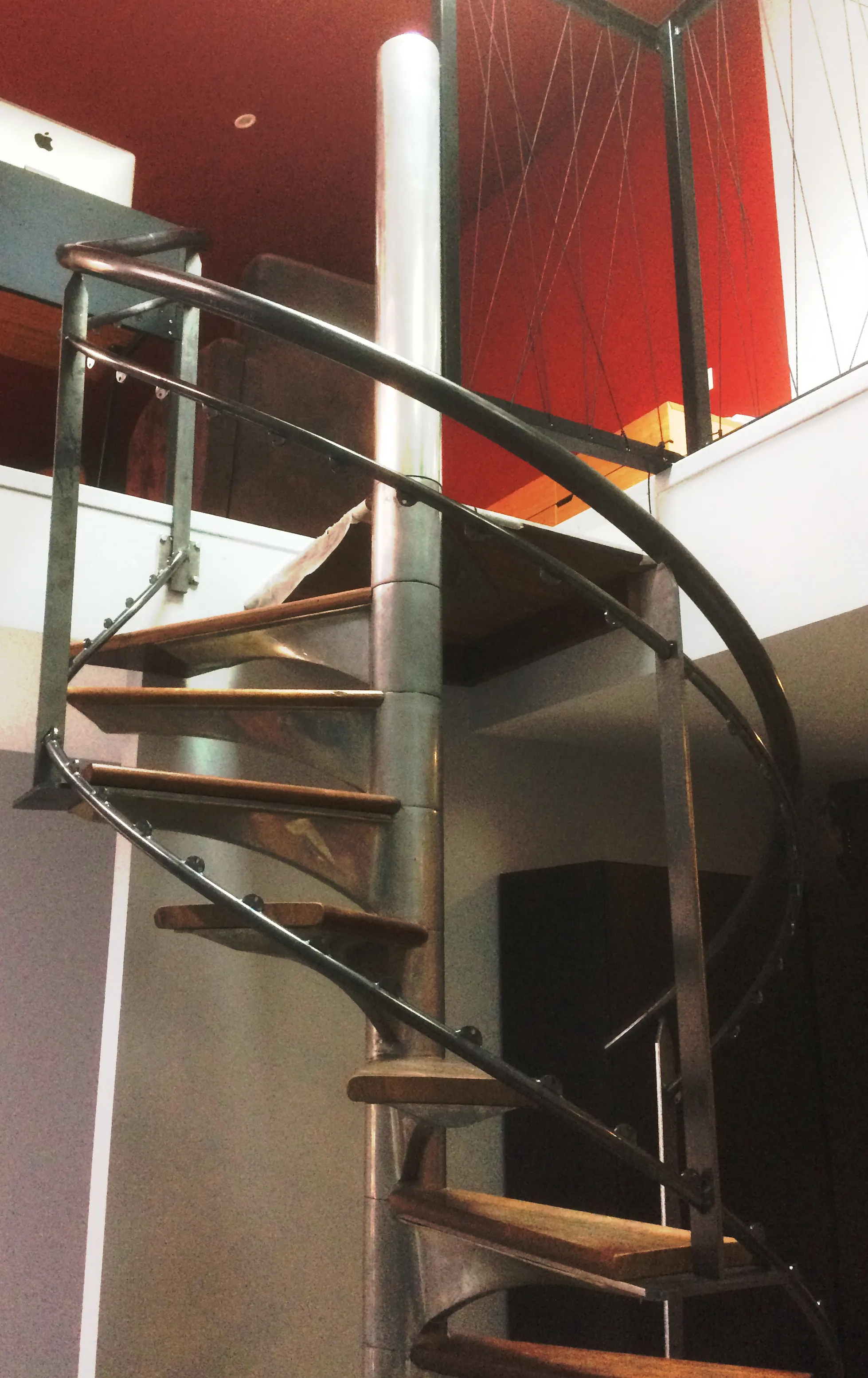 Escalier en colimaçon à marches de bois et main courante en acier, montant vers une mezzanine devant un mur rouge.