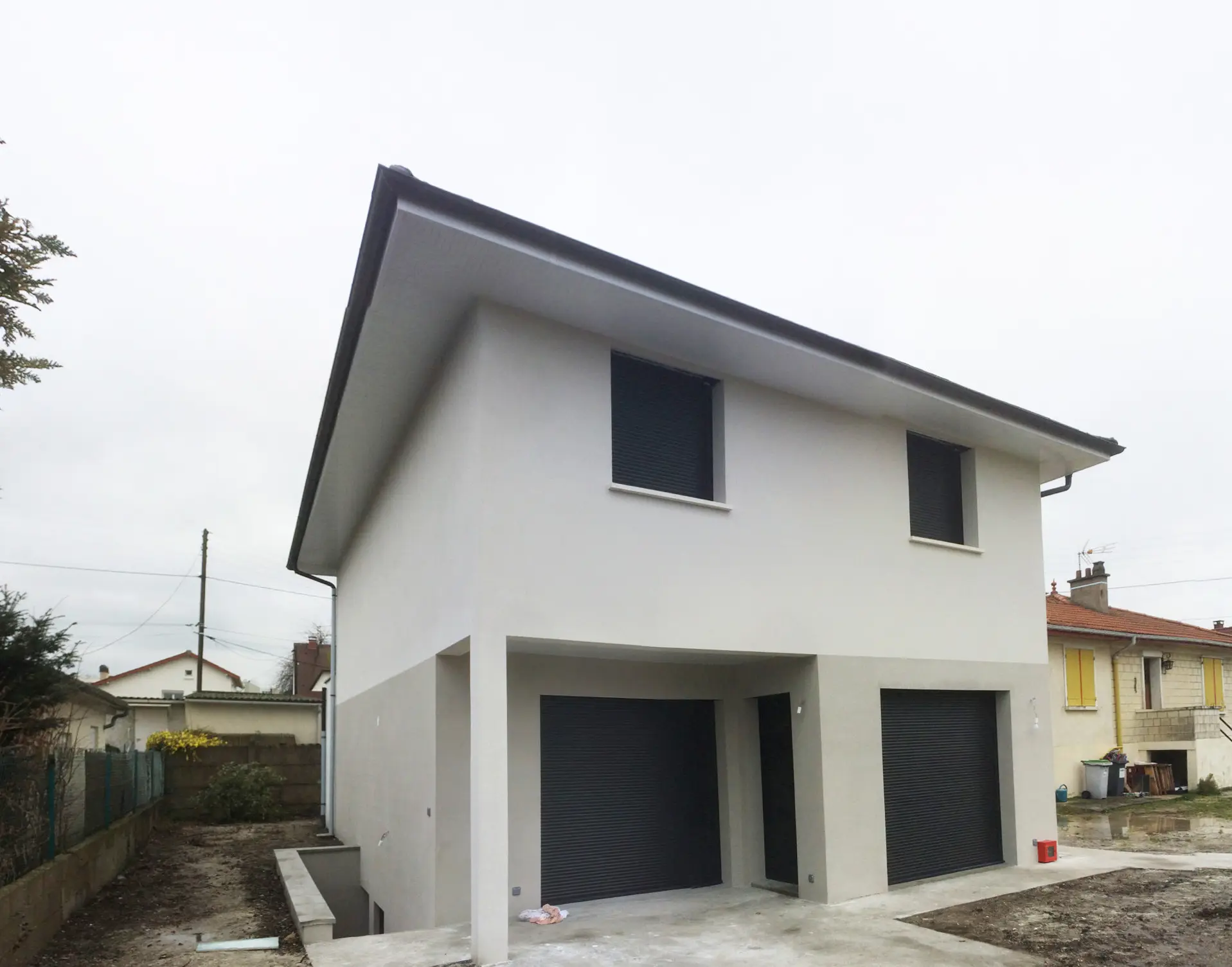 Maison enduite blanche à étage sous un toit à faible pente débordant, avec deux portes de garage au niveau inférieur et une terrasse en surplomb portée par un poteau.