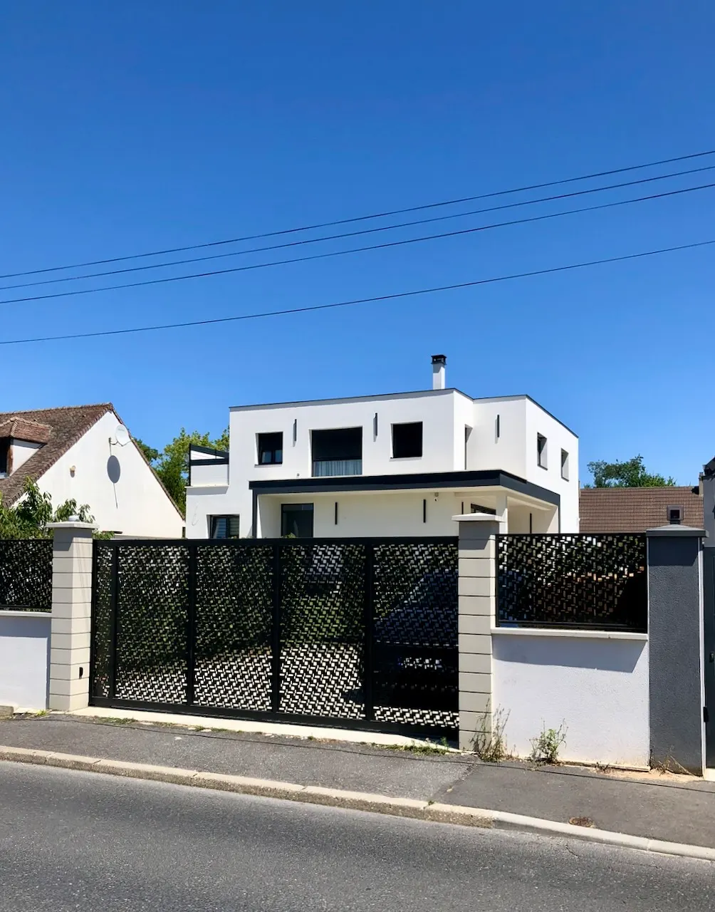 Maison blanche à toiture plate en fond de parcelle, vue à travers un portail ajouré entre deux piliers.