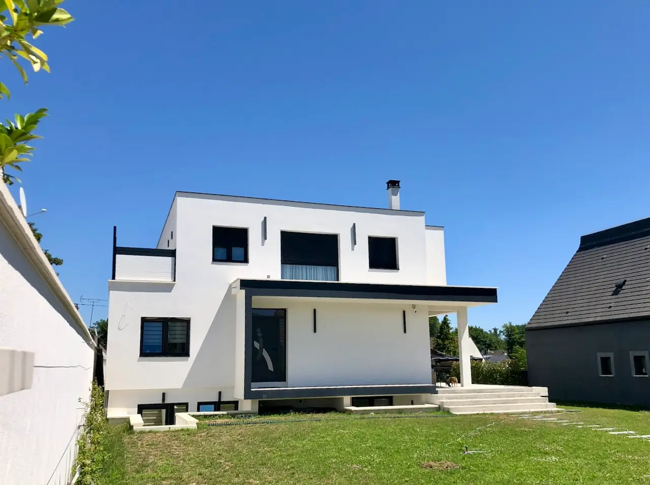 Maison contemporaine blanche à deux niveaux sur sous-sol, toiture plate, auvent sombre au rez-de-chaussée, pelouse au premier plan.