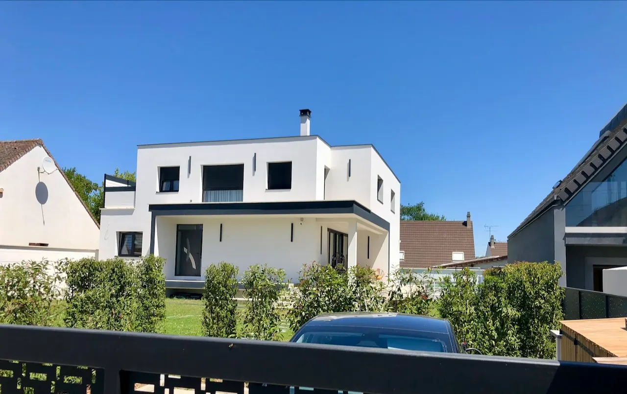 Maison blanche à toiture plate et larges baies, vue par-dessus une haie taillée, sous un ciel bleu.