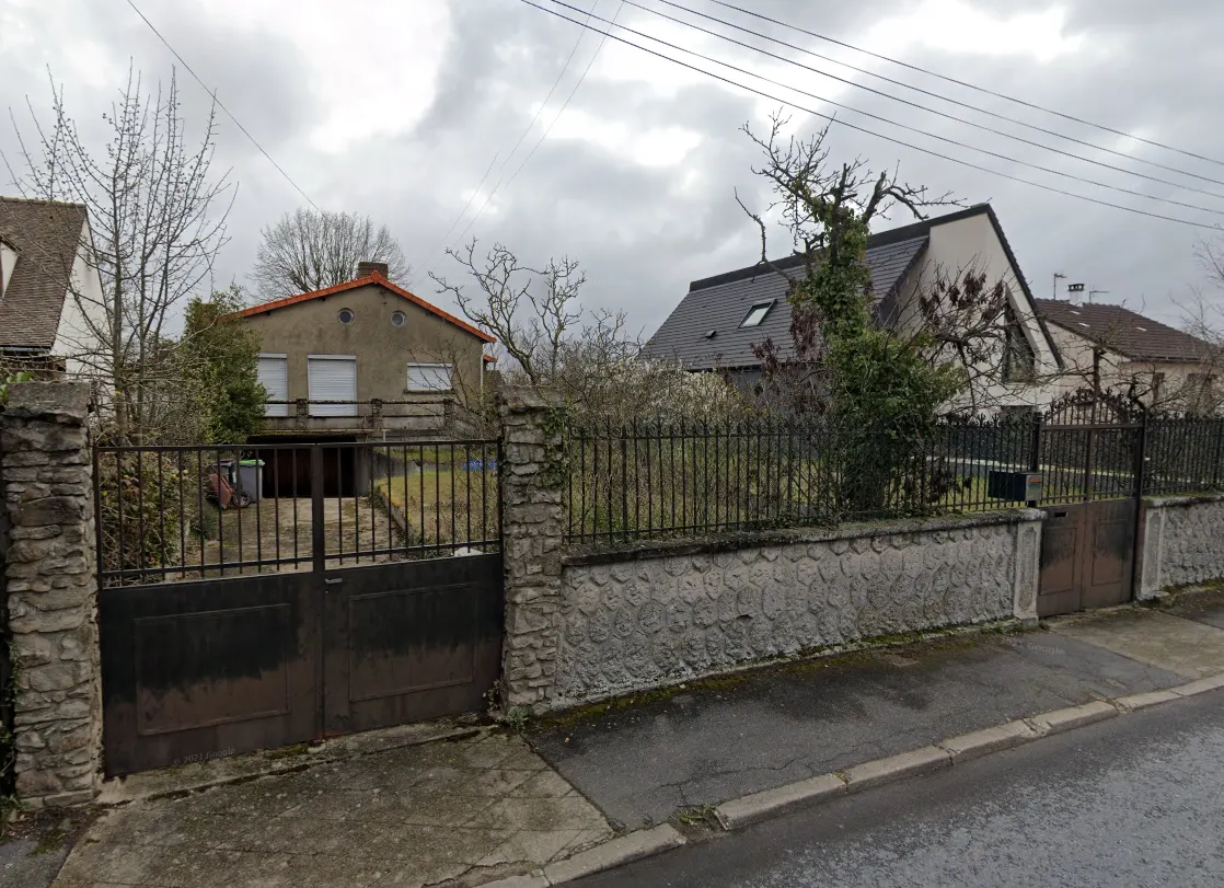 Maison ancienne à toit à deux pans, en retrait d'une rue, derrière un portail et un mur bas surmonté d’un grillage.