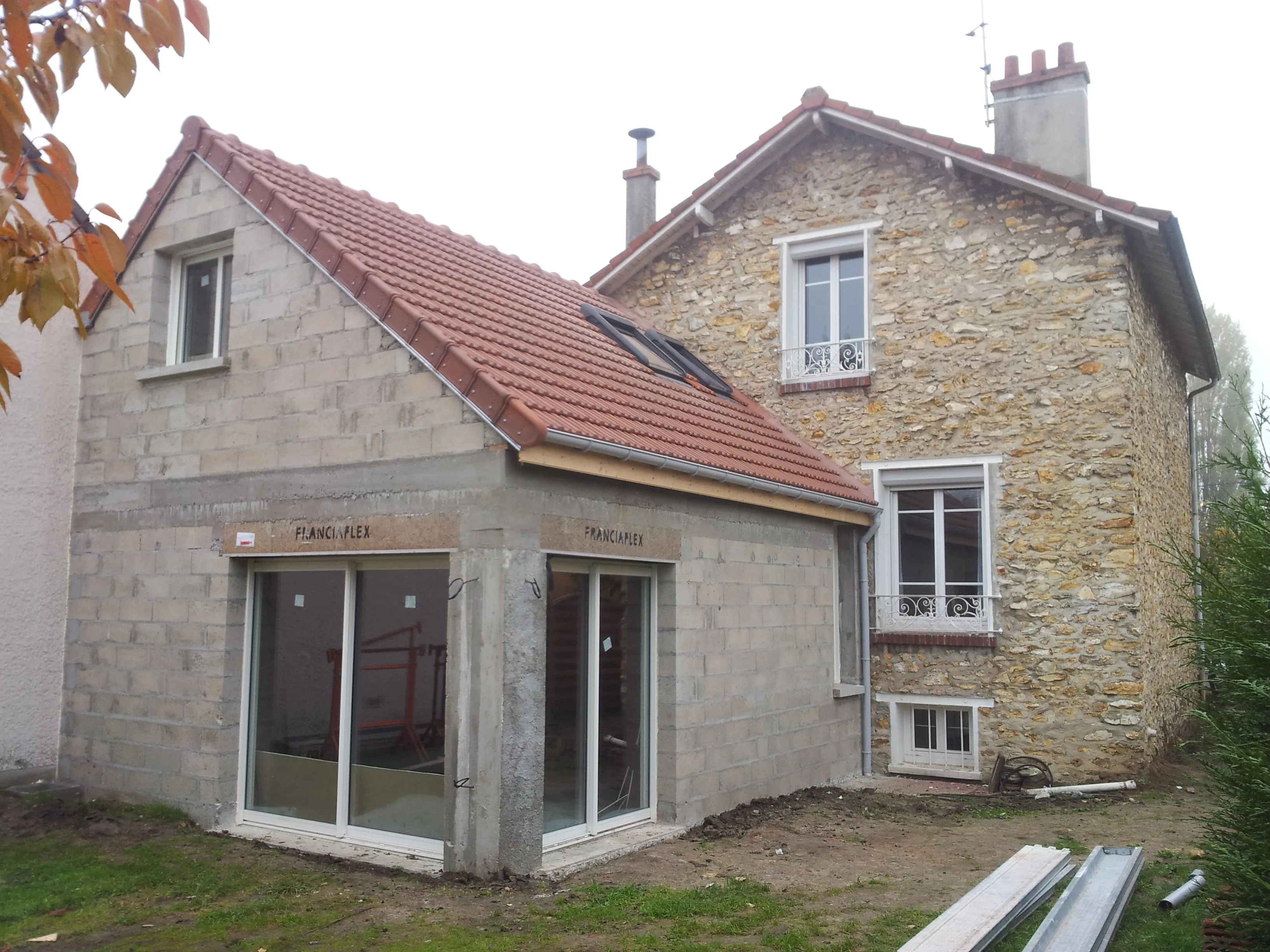 Extension en blocs de béton apparents sous une toiture de tuiles, avec deux baies vitrées posées, accolée à la maison en moellons.