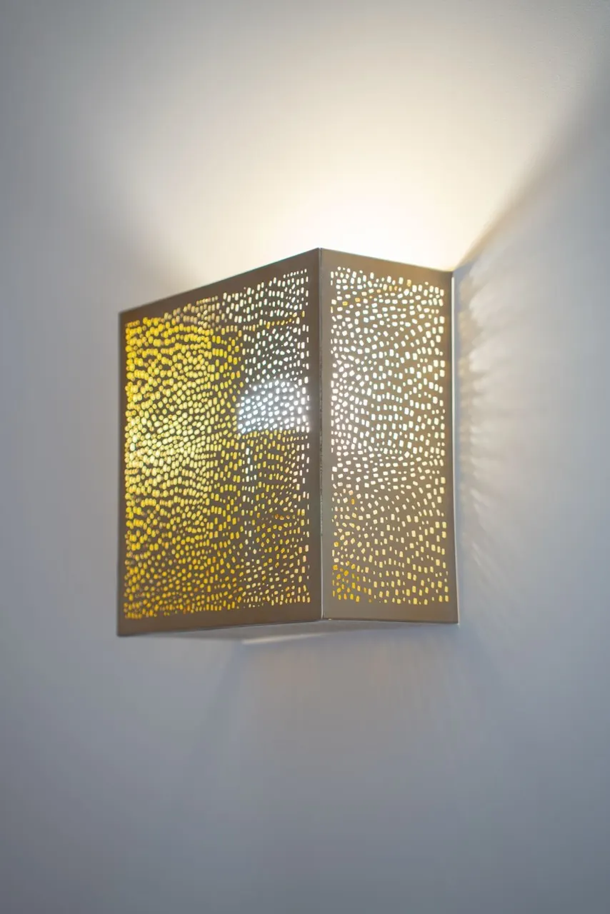 Applique murale cubique en métal doré perforé diffusant une lumière chaude sur un mur gris.