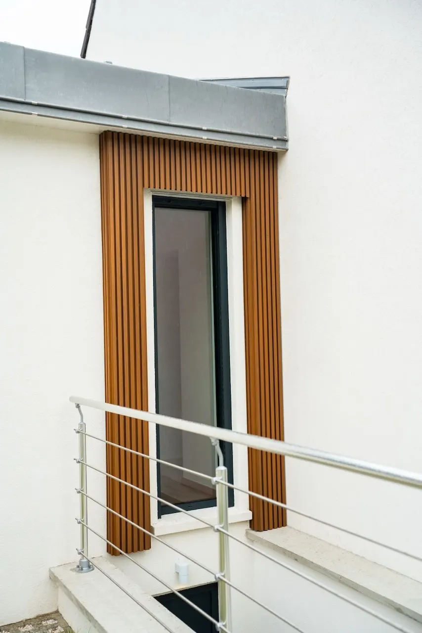 Porte vitrée encadrée de lames de bois verticales, sous un auvent métallique, avec un garde-corps à lisses horizontales au premier plan.
