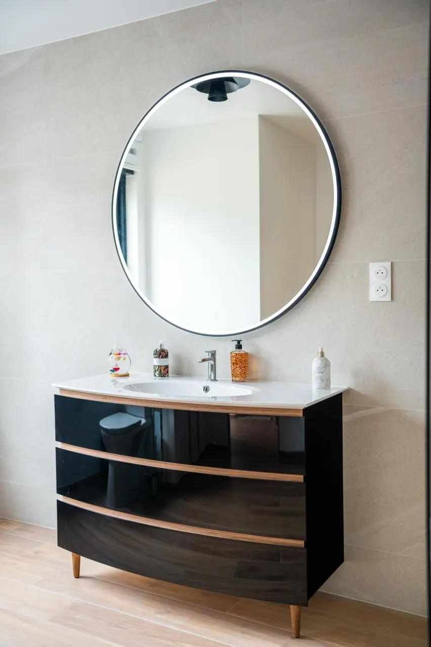 Meuble vasque laqué noir à façades galbées sous un miroir rond, devant un mur en grands carreaux clairs.