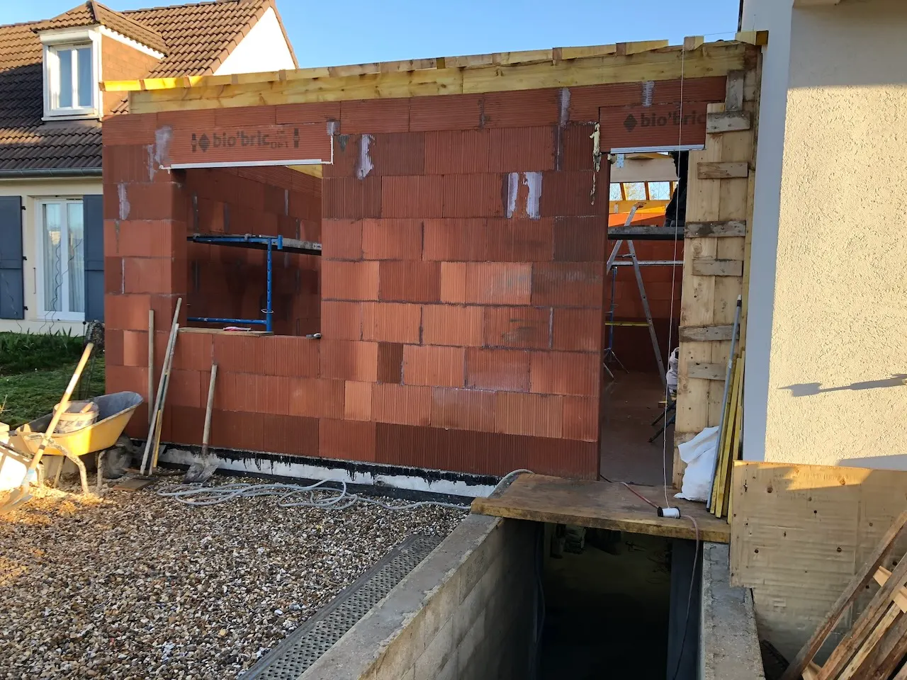 Murs en briques de terre cuite rouges en cours d'élévation, avec deux ouvertures réservées, accolés à une maison enduite, gravier et brouette au premier plan.